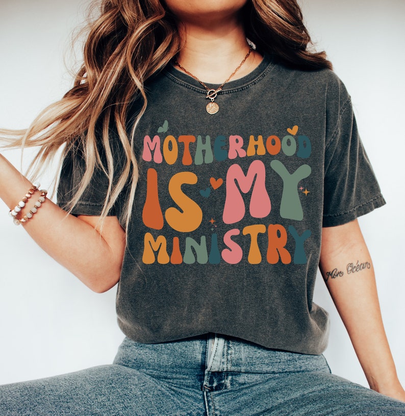 May include: A dark gray t-shirt with the text "Motherhood is my ministry" in colorful, retro-style lettering. The text is surrounded by small hearts and stars.