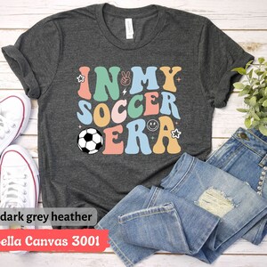 Może przedstawiać: Koszulka w kolorze ciemnoszary melanż Bella Canvas 3001 z napisem "In My Soccer Era" w kolorowych retro literach. W projekcie znajduje się piłka nożna.