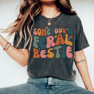 May include: A dark gray t-shirt with a colorful graphic that reads "Somebody's Feral Bestie".