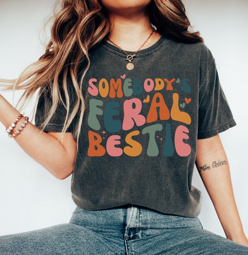 May include: A dark gray t-shirt with a colorful graphic that reads "Somebody's Feral Bestie".