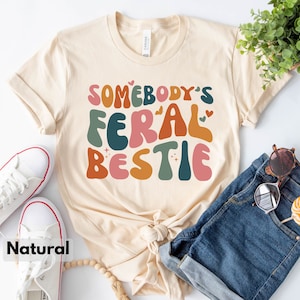 May include: A beige t-shirt with the text "Somebody's Feral Bestie" in colorful, retro-style lettering. The shirt is displayed on a white surface with a pair of white sneakers, a pair of blue denim shorts, and a wooden bead necklace.