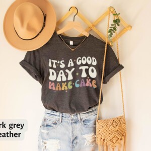 Peut inclure: T-shirt en jersey chin&eacute; gris fonc&eacute; avec le texte "It's a good day to make cake" en couleurs arc-en-ciel avec des &eacute;toiles. Le t-shirt est suspendu &agrave; un cintre en bois avec un sac en paille et un chapeau.