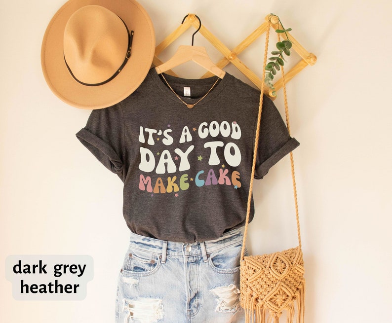Peut inclure: T-shirt en jersey chin&eacute; gris fonc&eacute; avec le texte "It's a good day to make cake" en couleurs arc-en-ciel avec des &eacute;toiles. Le t-shirt est suspendu &agrave; un cintre en bois avec un sac en paille et un chapeau.