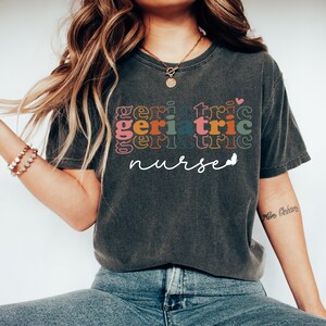 Może przedstawiać: Szary t-shirt z napisem "geriatric nurse" w kolorowej, nakładanej czcionce retro. Tekst jest w tęczy kolor&oacute;w, w tym r&oacute;żowy, pomarańczowy, ż&oacute;łty, zielony, niebieski i fioletowy.