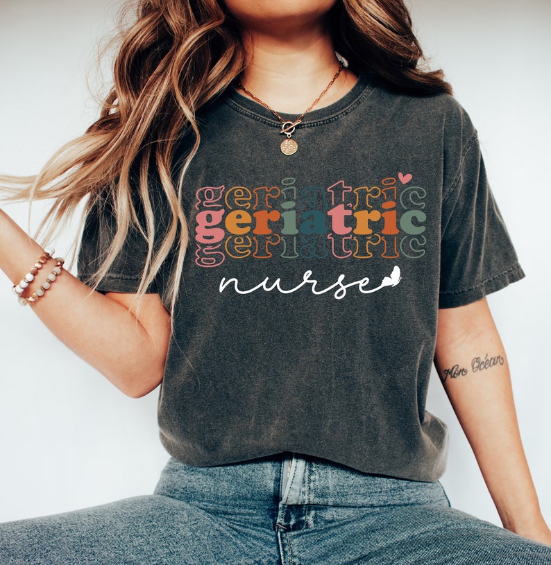 Może przedstawiać: Szary t-shirt z napisem "geriatric nurse" w kolorowej, nakładanej czcionce retro. Tekst jest w tęczy kolor&oacute;w, w tym r&oacute;żowy, pomarańczowy, ż&oacute;łty, zielony, niebieski i fioletowy.