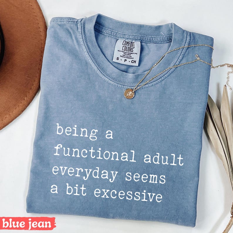 K&ouml;nnte beinhalten: Blaues Jeans-T-Shirt mit dem wei&szlig;en Text "being a functional adult everyday seems a bit excessive". Eine goldene Halskette mit Anh&auml;nger liegt &uuml;ber dem Shirt.
