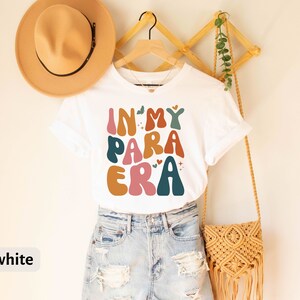 May include: White t-shirt with the text "In My Para Era" in colorful, retro-style lettering. The shirt is hanging on a wooden rack with a straw hat and a woven bag.