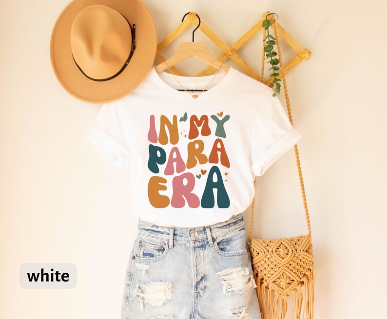May include: White t-shirt with the text "In My Para Era" in colorful, retro-style lettering. The shirt is hanging on a wooden rack with a straw hat and a woven bag.