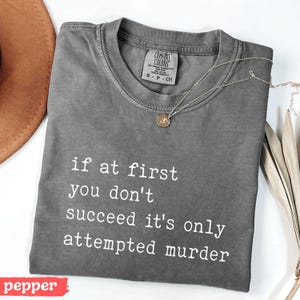 Op de afbeelding: Een grijs Comfort Colors T-shirt met de tekst "if at first you don't succeed it's only attempted murder" in het wit. Een gouden ketting met een hanger ligt op het shirt. Het woord "pepper" staat in een rode rechthoek.