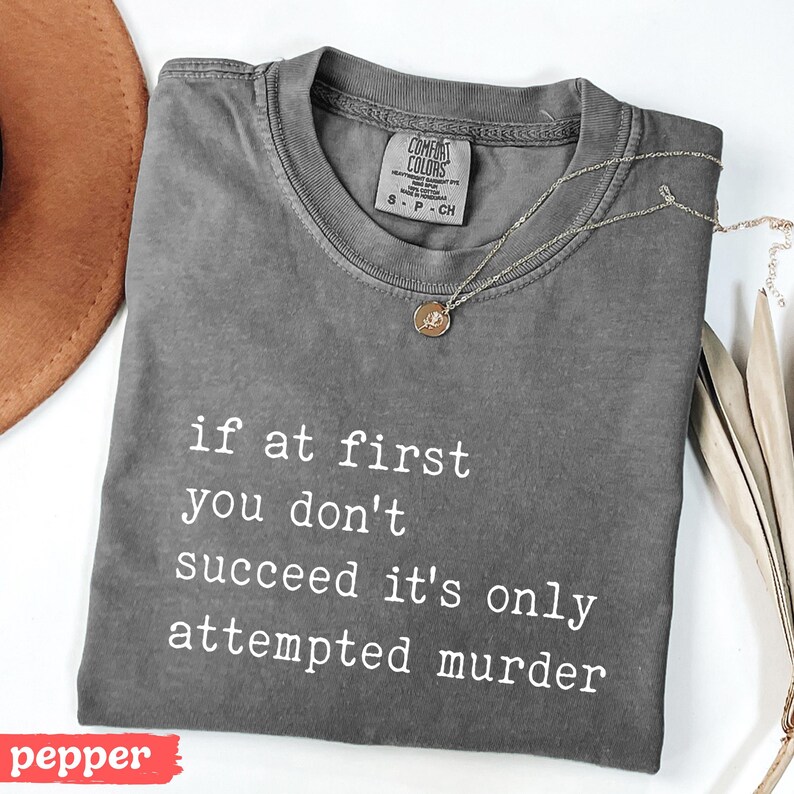 Op de afbeelding: Een grijs Comfort Colors T-shirt met de tekst "if at first you don't succeed it's only attempted murder" in het wit. Een gouden ketting met een hanger ligt op het shirt. Het woord "pepper" staat in een rode rechthoek.