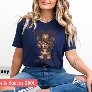 Puede incluir: Camiseta azul marino con un dise&ntilde;o de un perro salchicha con sombrero de vaquero. El texto "HOWDY DACHSHUND" est&aacute; encima y debajo del perro. La camiseta est&aacute; etiquetada como "Bella Canvas 3001".