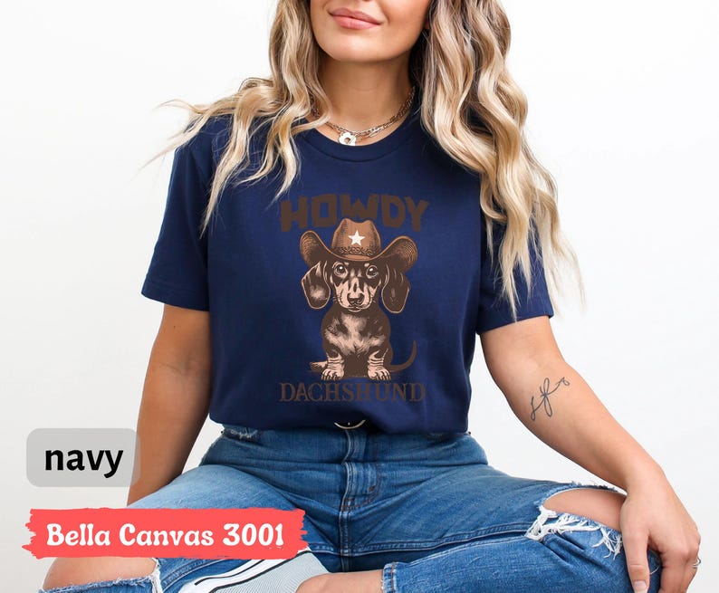 Puede incluir: Camiseta azul marino con un dise&ntilde;o de un perro salchicha con sombrero de vaquero. El texto "HOWDY DACHSHUND" est&aacute; encima y debajo del perro. La camiseta est&aacute; etiquetada como "Bella Canvas 3001".
