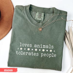 May include: A green t-shirt with the text "loves animals tolerates people" in white lettering with small white flowers between the words. The shirt is made by Comfort Colors and is in the color moss.