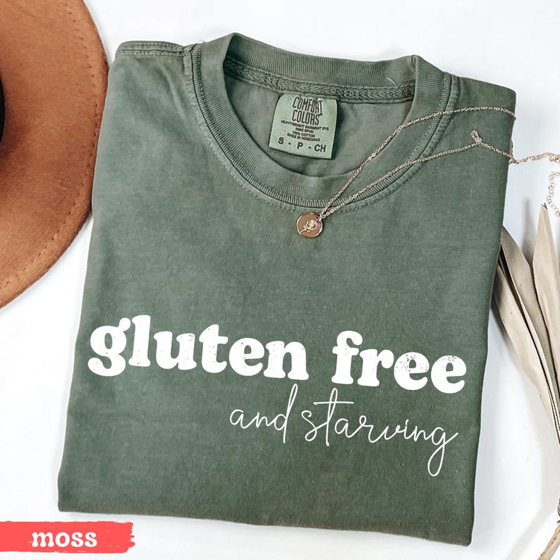 Puede incluir: Una camiseta verde musgo con las palabras "gluten free and starving" en blanco. La camiseta tiene cuello redondo y mangas cortas. Un collar dorado con un colgante est&aacute; sobre la camiseta.