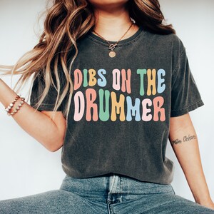 Puede incluir: Una camiseta gris oscuro con el texto "DIBS ON THE DRUMMER" en letras coloridas y onduladas. Las letras son en tonos de rosa, amarillo, verde y azul.