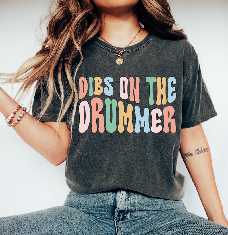 Puede incluir: Una camiseta gris oscuro con el texto "DIBS ON THE DRUMMER" en letras coloridas y onduladas. Las letras son en tonos de rosa, amarillo, verde y azul.