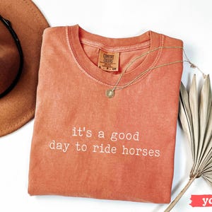 May include: A coral-colored t-shirt with the text "it's a good day to ride horses" in white embroidery.