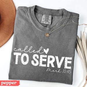 Pu&ograve; includere: Una maglietta grigia Comfort Colors con le parole "called TO SERVE" in corsivo bianco e lettere maiuscole, con "Mark 10:45" sotto. Una collana d'oro con un ciondolo poggia sulla maglietta. Un cappello marrone &egrave; visibile in alto a sinistra.