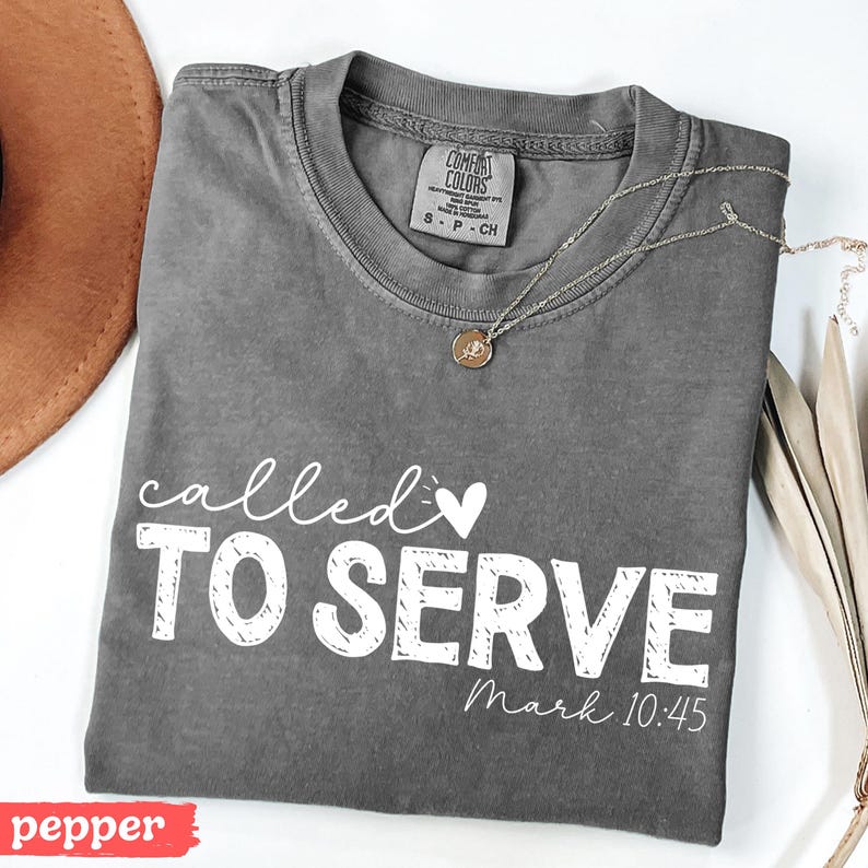 Pu&ograve; includere: Una maglietta grigia Comfort Colors con le parole "called TO SERVE" in corsivo bianco e lettere maiuscole, con "Mark 10:45" sotto. Una collana d'oro con un ciondolo poggia sulla maglietta. Un cappello marrone &egrave; visibile in alto a sinistra.