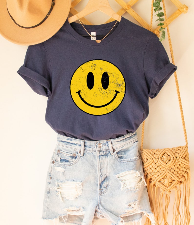 May include: A navy blue t-shirt featuring a yellow smiley face graphic. The smiley face has a distressed look, giving it a vintage feel.