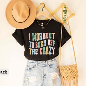 May include: Black t-shirt with the text "I workout to burn off the crazy" in colorful letters. The shirt is hanging on a wooden rack with a straw hat and a woven bag.