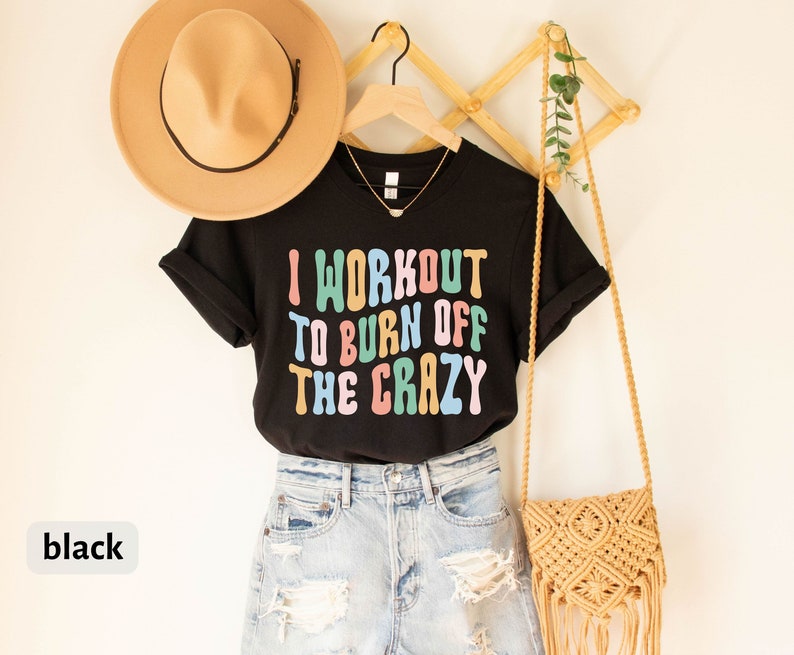 May include: Black t-shirt with the text "I workout to burn off the crazy" in colorful letters. The shirt is hanging on a wooden rack with a straw hat and a woven bag.