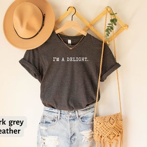 May include: Dark grey heather V-neck t-shirt with the text "I'm a Delight." printed in white. The shirt is hanging on a wooden hanger with a straw bag and a hat.