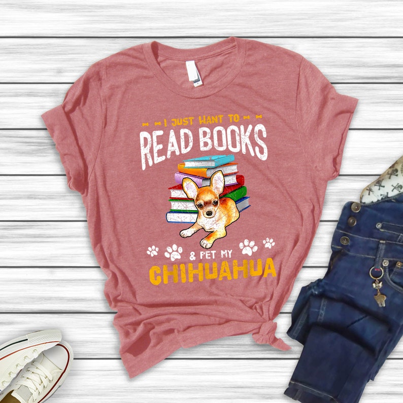 May include: A pink t-shirt with the text "I just want to read books & pet my chihuahua" in a distressed font. A chihuahua is sitting on top of a stack of books. The chihuahua is brown and white with floppy ears.
