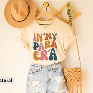 May include: A beige t-shirt with the text "In My Para Era" in colorful, retro-style lettering. The shirt is hanging on a wooden hanger and has a natural color.