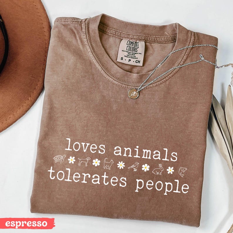 May include: A brown t-shirt with the text "loves animals tolerates people" in white. The text is surrounded by small white animal silhouettes and white flowers.