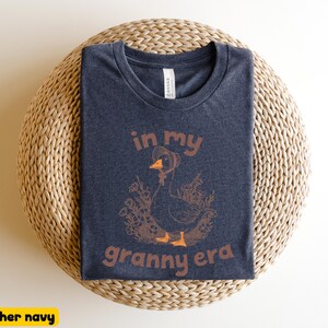 May include: Heather navy t-shirt with a graphic of a goose wearing a hat, surrounded by flowers, and the text "in my granny era" in brown. The shirt is folded on a woven, circular surface.