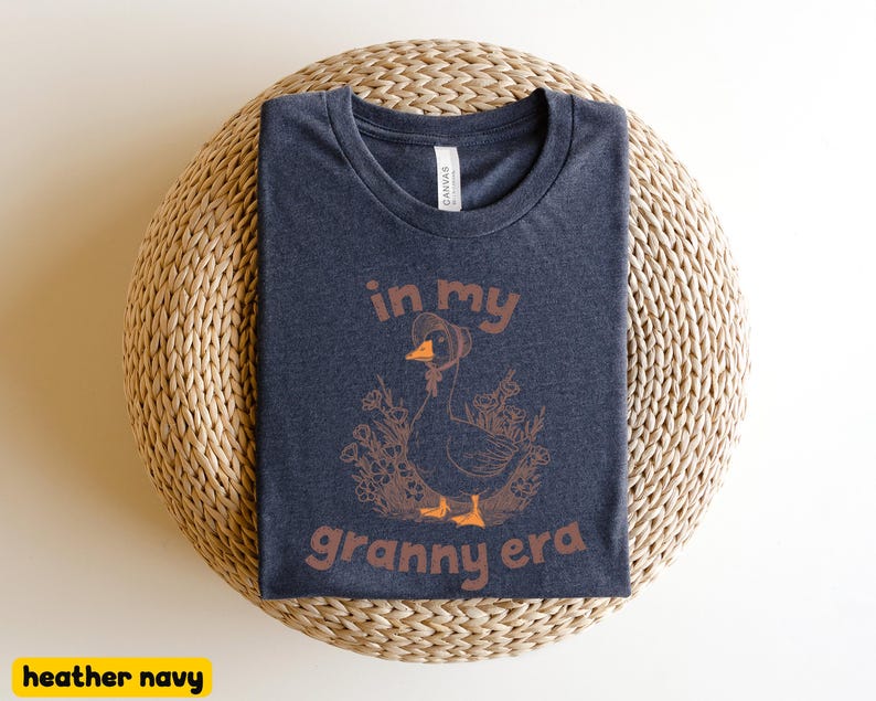May include: Heather navy t-shirt with a graphic of a goose wearing a hat, surrounded by flowers, and the text "in my granny era" in brown. The shirt is folded on a woven, circular surface.