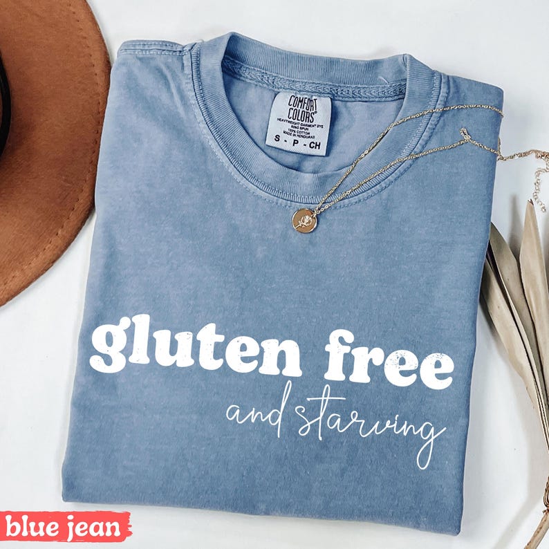 Puede incluir: Camiseta azul vaquero con las palabras blancas "gluten free and starving". La camiseta tiene cuello redondo y mangas cortas. Un collar dorado con un colgante est&aacute; sobre la camiseta.
