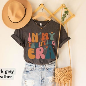 May include: Dark grey heather t-shirt with the text "IN MY THRIFTING ERA" in retro-style lettering. The shirt is hanging on a wooden hanger, with a tan hat and a macrame purse visible in the background.