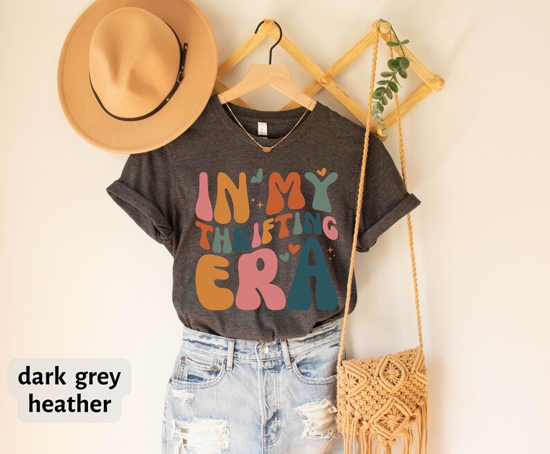May include: Dark grey heather t-shirt with the text "IN MY THRIFTING ERA" in retro-style lettering. The shirt is hanging on a wooden hanger, with a tan hat and a macrame purse visible in the background.