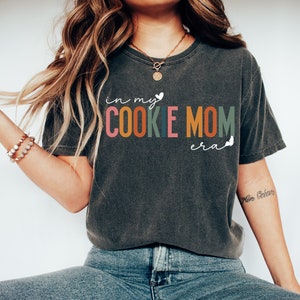 May include: A dark gray t-shirt with the text "in my cookie mom era" in colorful letters. The text is in a playful, handwritten font.