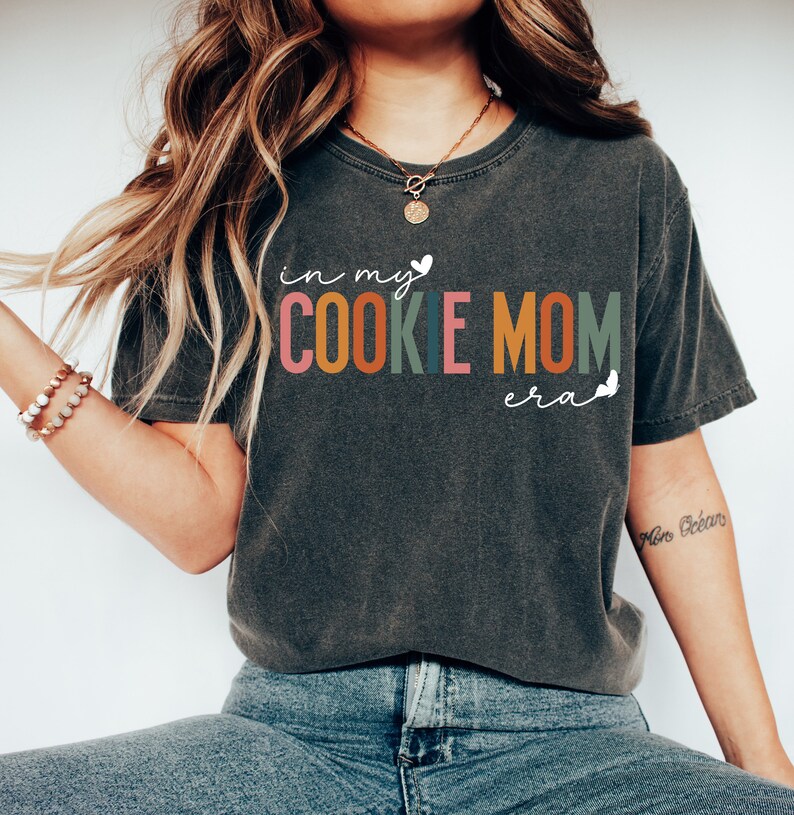 May include: A dark gray t-shirt with the text "in my cookie mom era" in colorful letters. The text is in a playful, handwritten font.