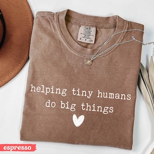May include: Espresso-colored t-shirt with the phrase "helping tiny humans do big things" in white, along with a small white heart graphic. A silver necklace with a pendant is draped across the shirt. The shirt has a crew neck and a tag that says "Comfort Colors".