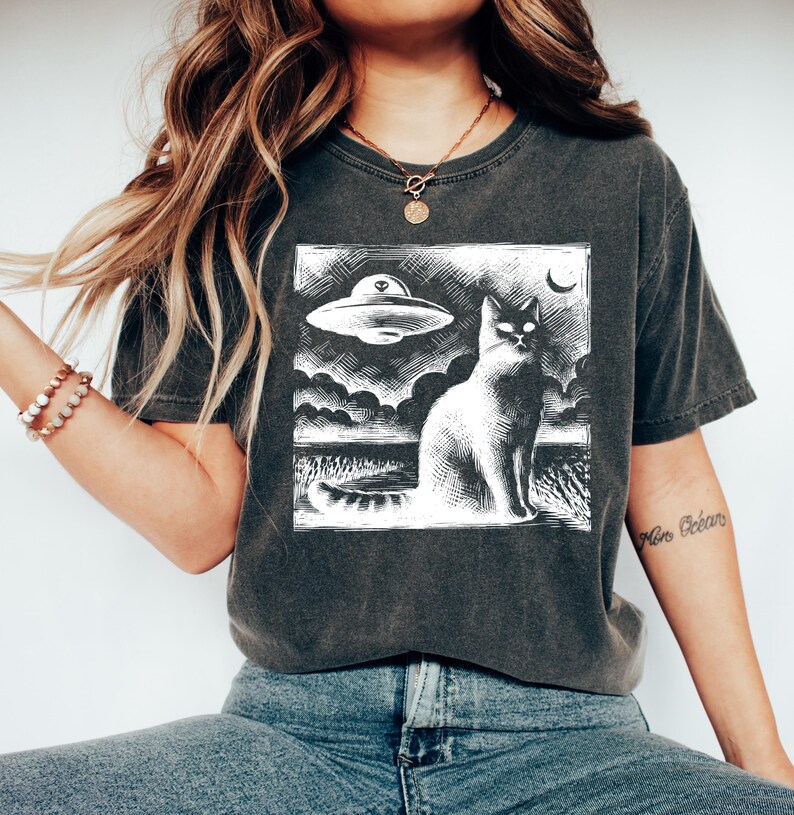 May include: A dark grey t-shirt with a white graphic print of a cat looking up at a UFO. The text "Moon Creatures" is printed on the right sleeve.