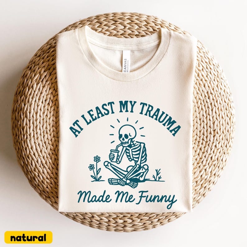 K&ouml;nnte beinhalten: Cremefarbenes T-Shirt mit dem Text "AT LEAST MY TRAUMA Made Me Funny" in T&uuml;rkis. Die Grafik zeigt ein Skelett, das aus einer Tasse trinkt, mit Blumen und Gras. Das Shirt ist aus weichem Material.