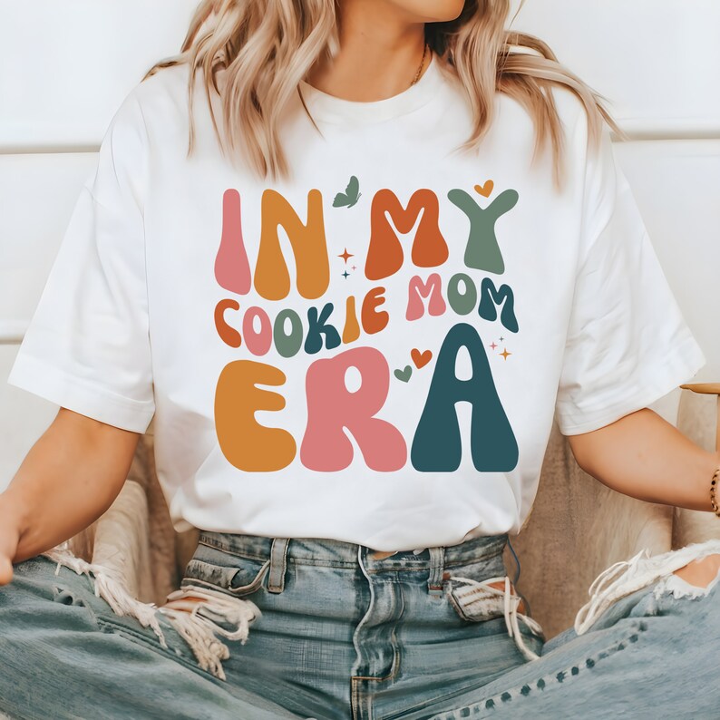 May include: White t-shirt with a colorful graphic that says "In My Cookie Mom Era". The graphic is in a retro style with rounded letters and hearts.