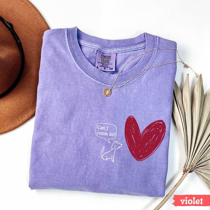 May include: A violet Comfort Colors t-shirt with a crew neck. The shirt features a red heart graphic and a white cartoon dog with the text "Can I come in?" A gold necklace is draped across the shirt.