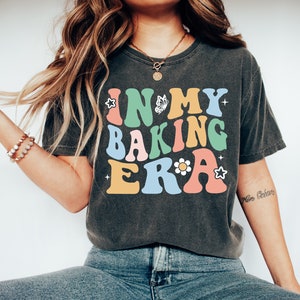 May include: A dark grey t-shirt with the text "IN MY BAKING ERA" in colorful retro-style lettering. The text is decorated with a butterfly, a flower, and stars.