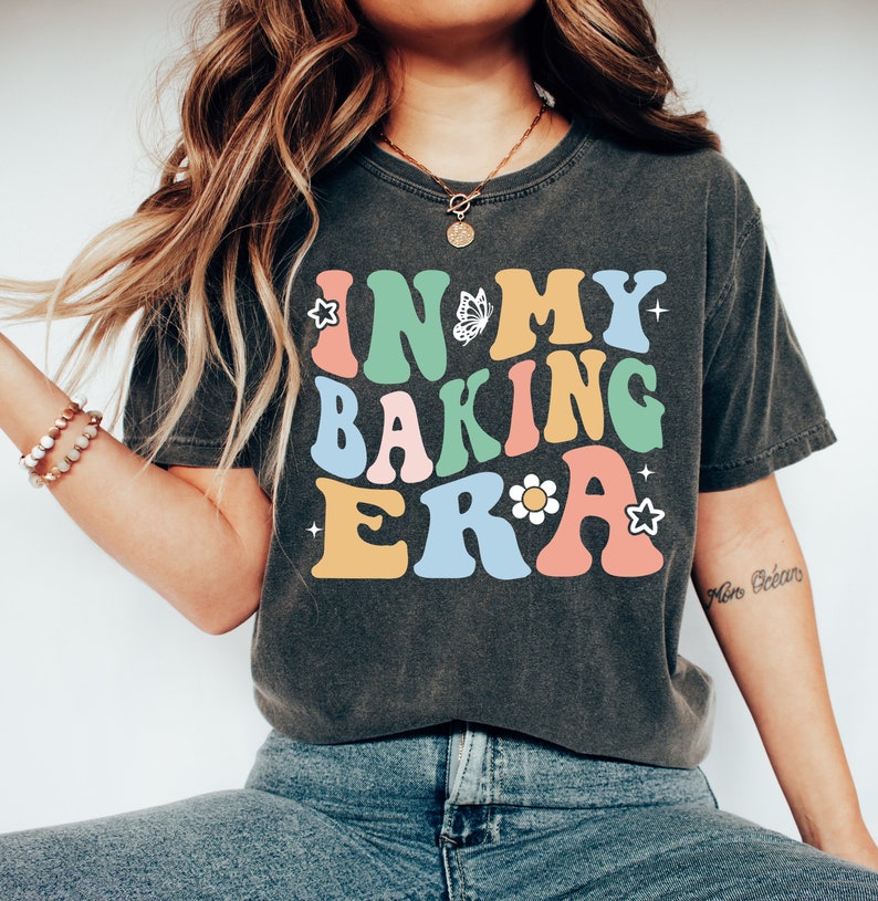 May include: A dark grey t-shirt with the text "IN MY BAKING ERA" in colorful retro-style lettering. The text is decorated with a butterfly, a flower, and stars.