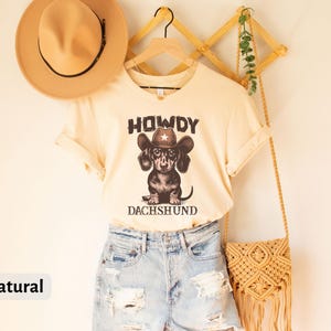 Puede incluir: Una camiseta color crema con el texto "HOWDY DACHSHUND" encima y debajo de un gr&aacute;fico de un perro salchicha con un sombrero de vaquero. La camiseta se combina con pantalones cortos de mezclilla y un sombrero color canela. Una bolsa tejida cuelga a la derecha.