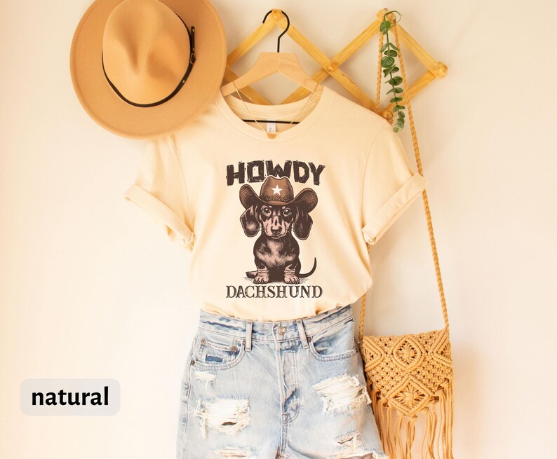 Puede incluir: Una camiseta color crema con el texto "HOWDY DACHSHUND" encima y debajo de un gr&aacute;fico de un perro salchicha con un sombrero de vaquero. La camiseta se combina con pantalones cortos de mezclilla y un sombrero color canela. Una bolsa tejida cuelga a la derecha.
