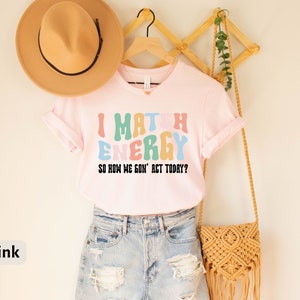 May include: Pink t-shirt with the text "I Match Energy So How We Gon' Act Today?" in colorful, retro-style letters.