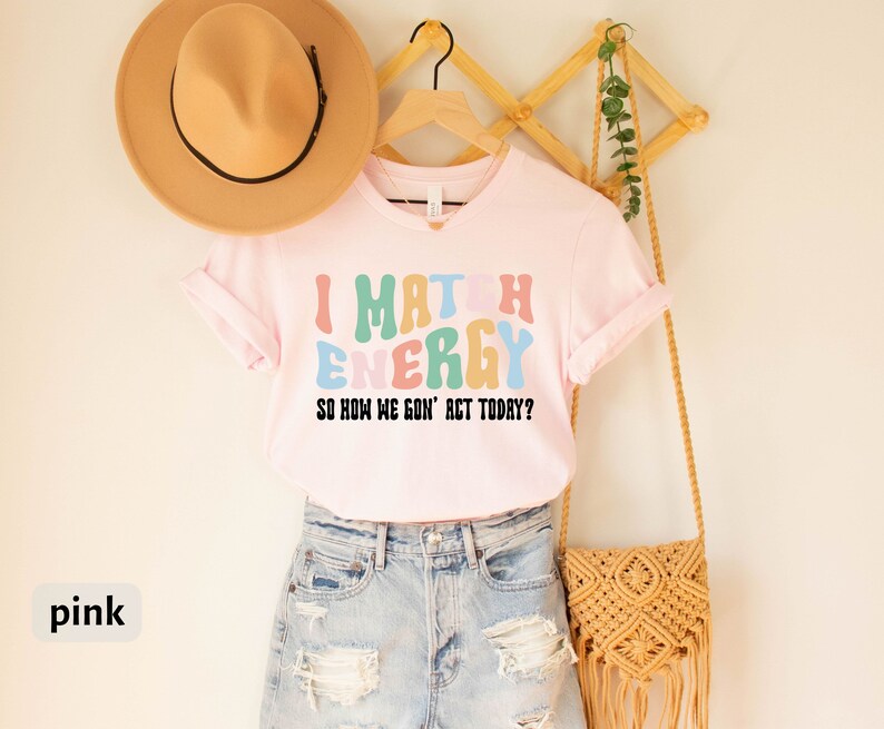 May include: Pink t-shirt with the text "I Match Energy So How We Gon' Act Today?" in colorful, retro-style letters.