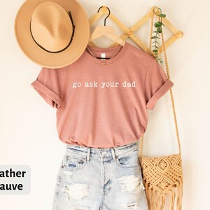 May include: Heather mauve t-shirt with the text "go ask your dad" in white lettering. The t-shirt is hanging on a wooden hanger with a straw bag and a potted plant.