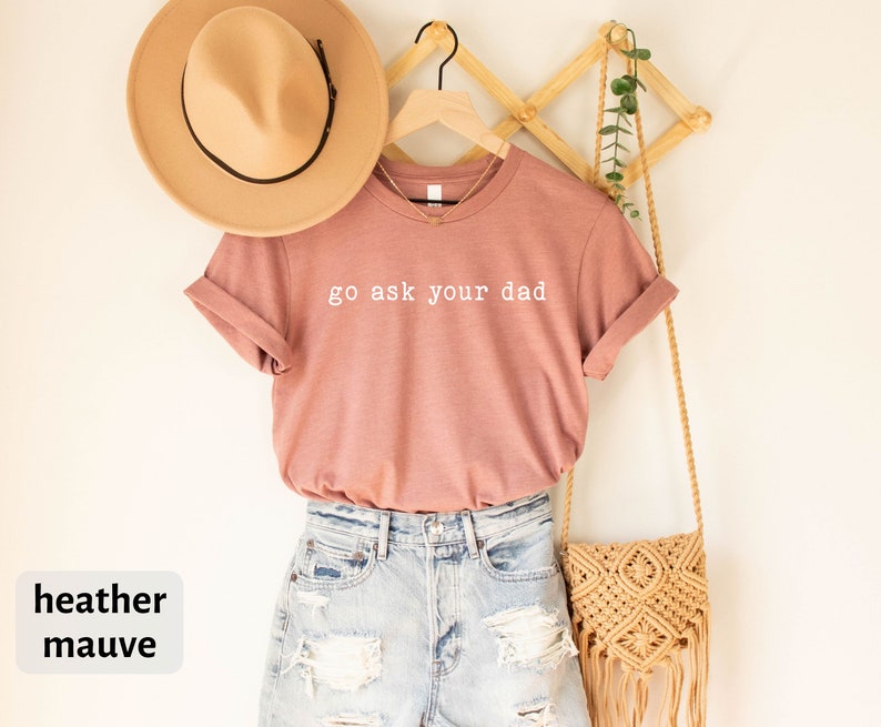 May include: Heather mauve t-shirt with the text "go ask your dad" in white lettering. The t-shirt is hanging on a wooden hanger with a straw bag and a potted plant.